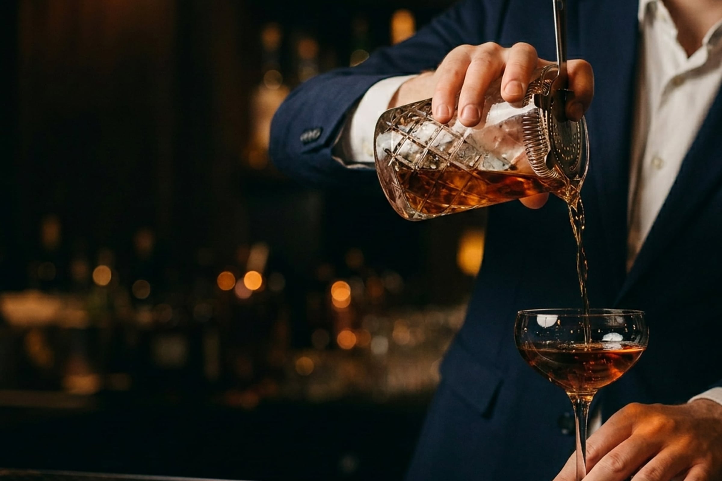 Bartender in a navy jacket pouring a cocktail into a coupe